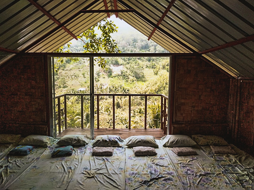 A bright loft area features a spacious sleeping arrangement with multiple pillows arranged on a large mattress. A wide glass door opens to a balcony, revealing a scenic view of lush greenery and the surrounding landscape.