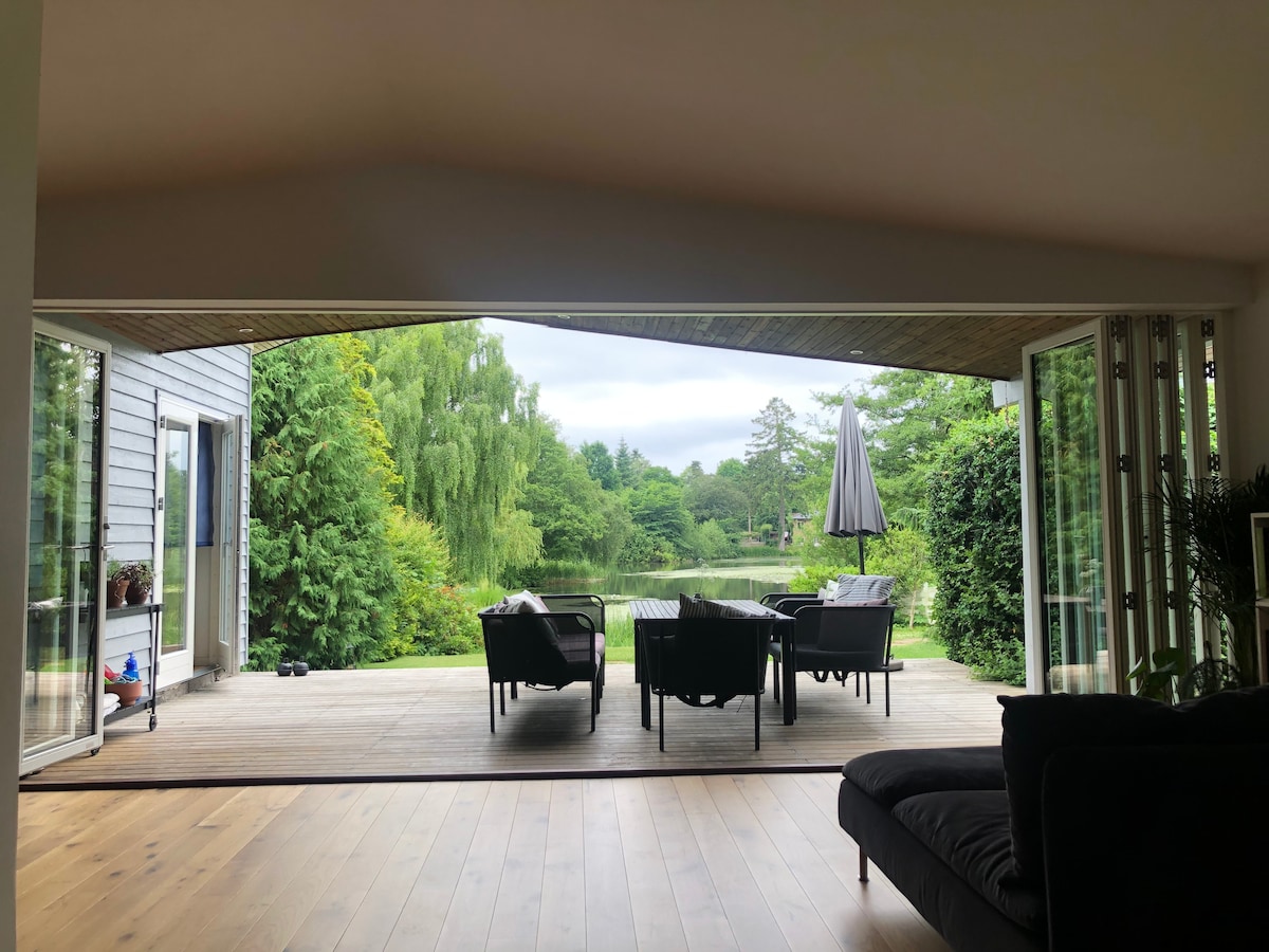 A spacious patio area is framed by large, folding glass doors, providing an unobstructed view of the serene lake. Comfortable seating arrangements include chairs and a table, enhanced by surrounding greenery and a shaded umbrella, creating a calm outdoor retreat.