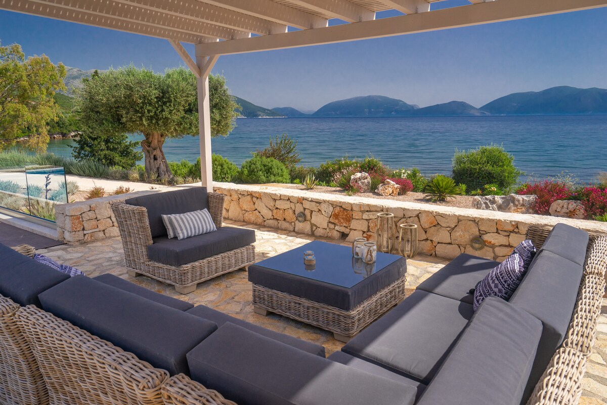 A spacious outdoor living area features comfortable seating arranged around a coffee table. A panoramic view of the Ionian Sea and lush greenery is visible in the background, creating a serene setting under a pergola.