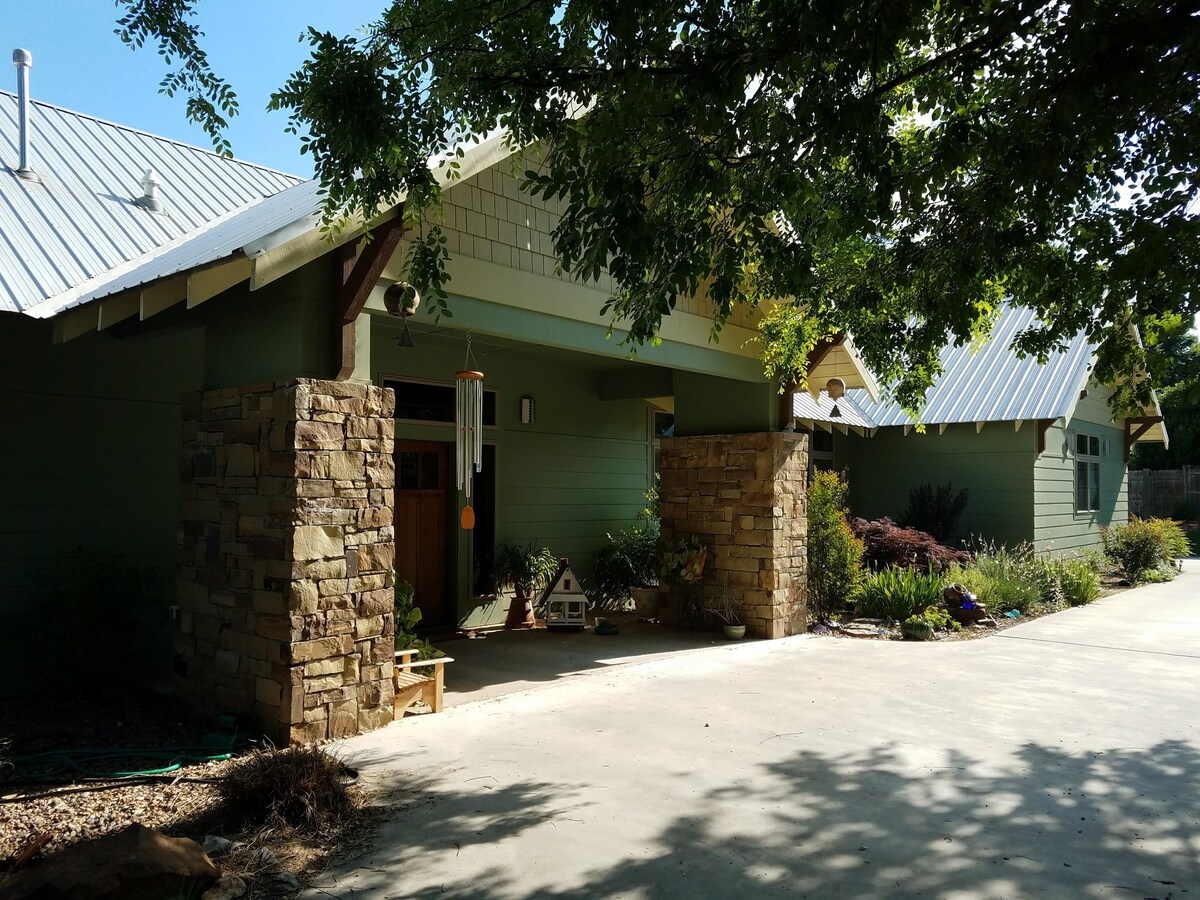 The exterior view of the bungalow showcases a blend of stone and green siding. A covered porch is framed by decorative pillars, with a wind chime hanging nearby. Lush landscaping, including shrubs and flowers, complements the home against a bright, sunlit sky.