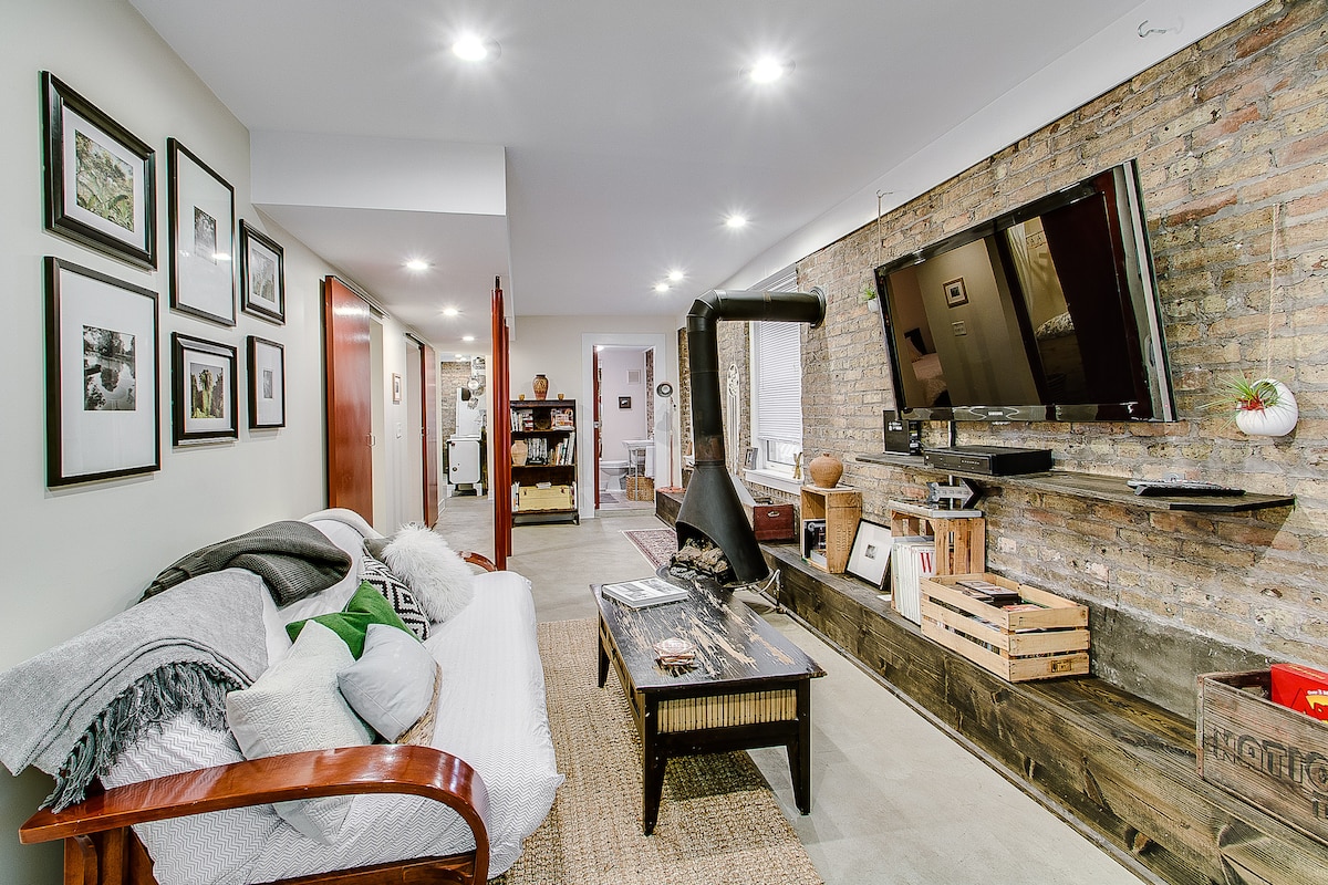 A cozy living area features an exposed brick wall, offering a rustic touch. A comfortable couch is paired with a wooden coffee table, while artwork hangs on the walls. A television is mounted above a rustic shelf, and soft lighting enhances the warm ambiance.