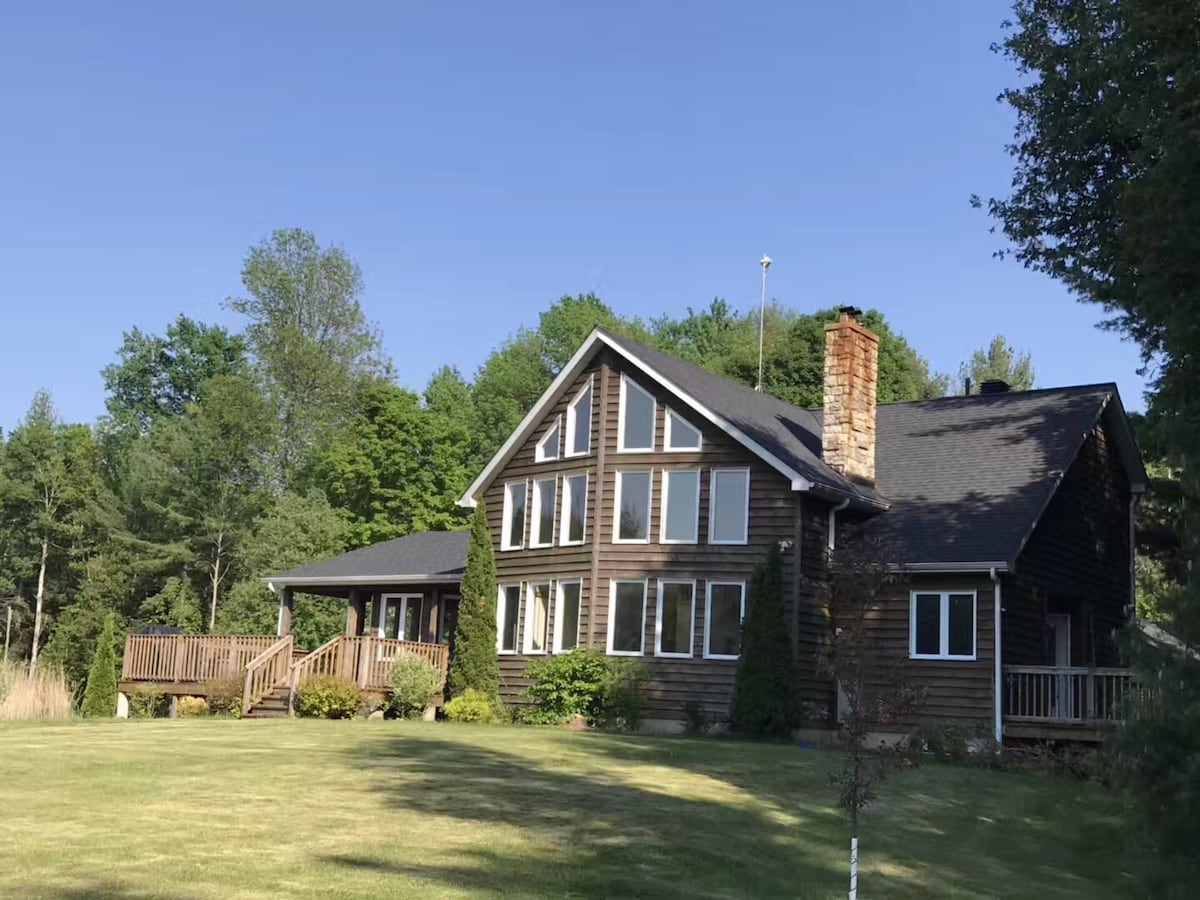 The custom-built country residence is showcased with a wide lawn in the foreground. The exterior features natural wood siding and multiple large windows, providing ample light. A stone chimney rises from the side of the building, surrounded by trees that enhance the secluded atmosphere.
