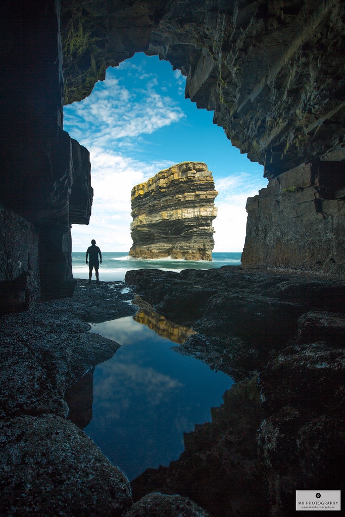 A rocky cave entrance frames a tall sea stack emerging from the water. The reflection of the stack is visible in a shallow pool at the cave's floor. The sky above features scattered clouds that illuminate the scene.
