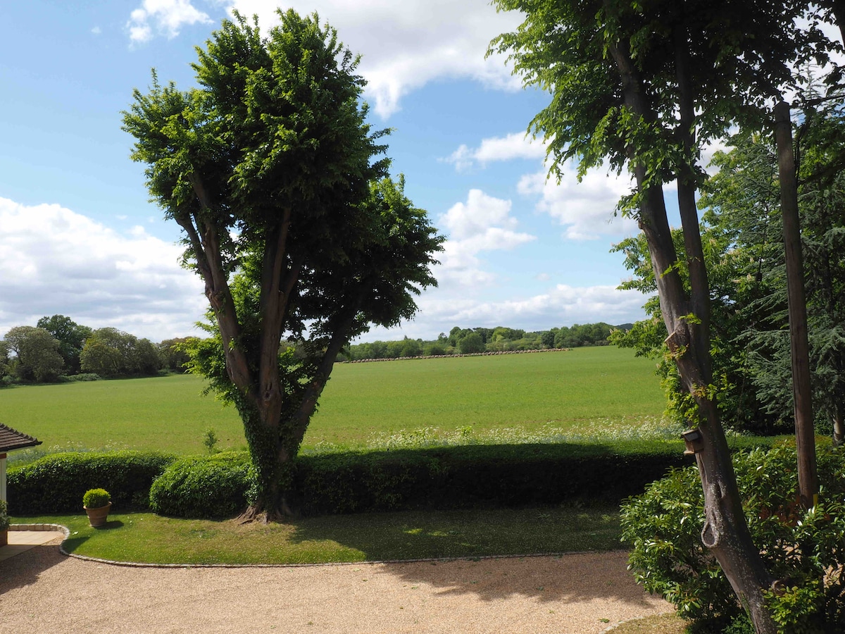 A scenic view showcases expansive green fields bordered by hedges and trees, under a partly cloudy sky. The image highlights the tranquility of the surrounding countryside, providing a relaxed atmosphere for guests.