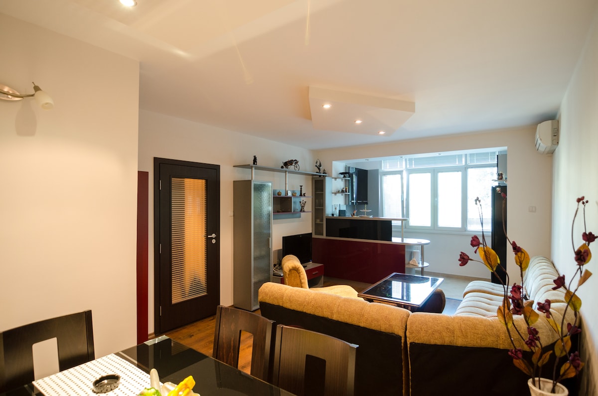 The open living area features a spacious layout with a comfortable sofa facing a sleek kitchen. A dining table is positioned nearby, complemented by warm lighting and a large window that offers natural light to the space.