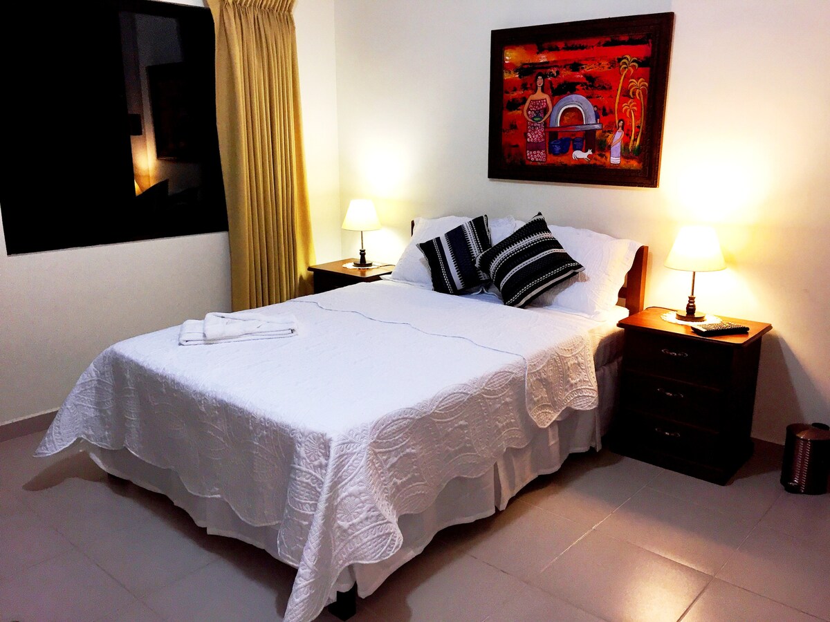 A well-appointed sleeping area features a spacious bed dressed in white linens, complemented by decorative pillows. Nightstands with lamps provide soft lighting, while a colorful piece of wall art adds character to the room. Natural light filters in through a nearby window covered by golden curtains.