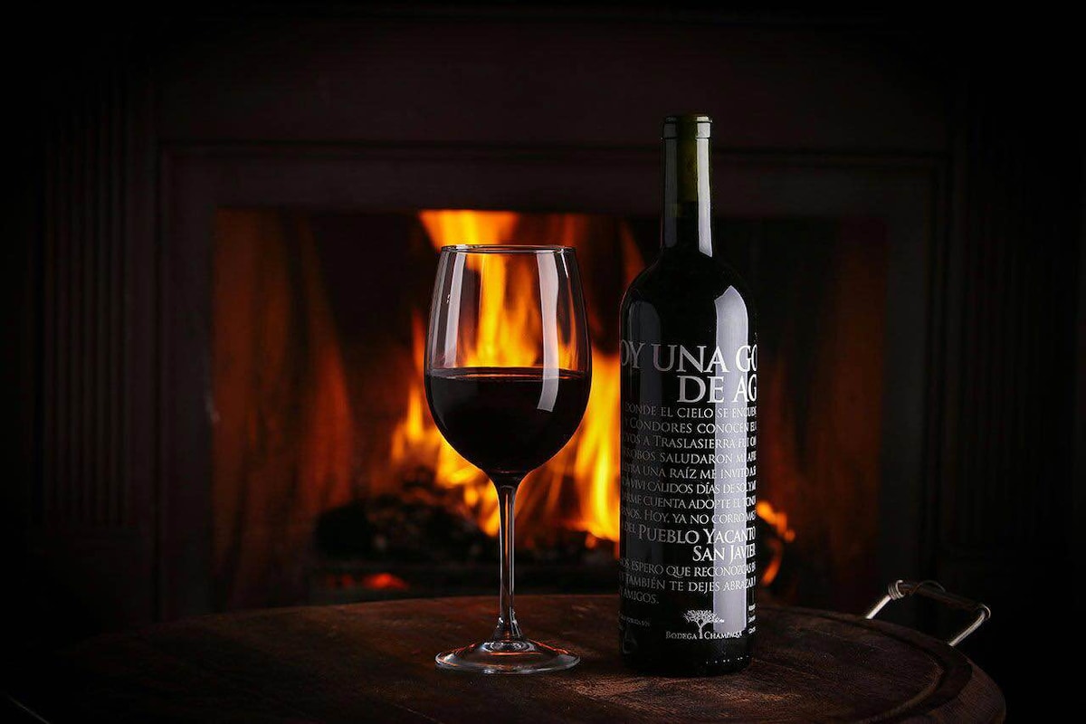 A glass of red wine is positioned beside a full wine bottle, both set against a rustic fireplace. The flames flicker warmly in the background, creating an inviting atmosphere with a soft glow illuminating the surroundings.