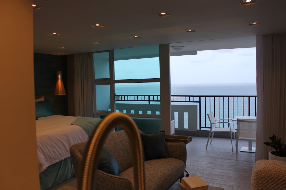 An open living space is featured, showcasing a spacious bed and modern seating. Large glass doors lead to a balcony overlooking the ocean. A small table with chairs is positioned on the balcony, allowing for coastal views and natural light to fill the room.