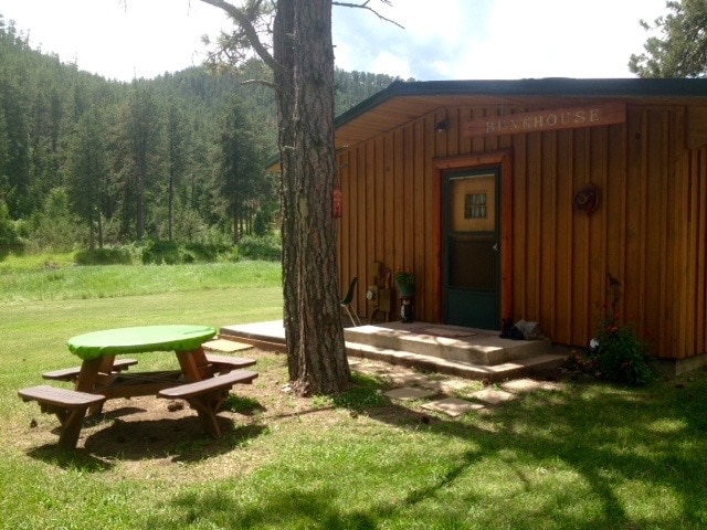 A cabin exterior is shown, featuring wooden paneling and a welcoming entrance. Outdoor seating is provided by a circular picnic table in vibrant green and two benches. Surrounding grassy areas offer ample space for play, with a serene backdrop of trees and hills.