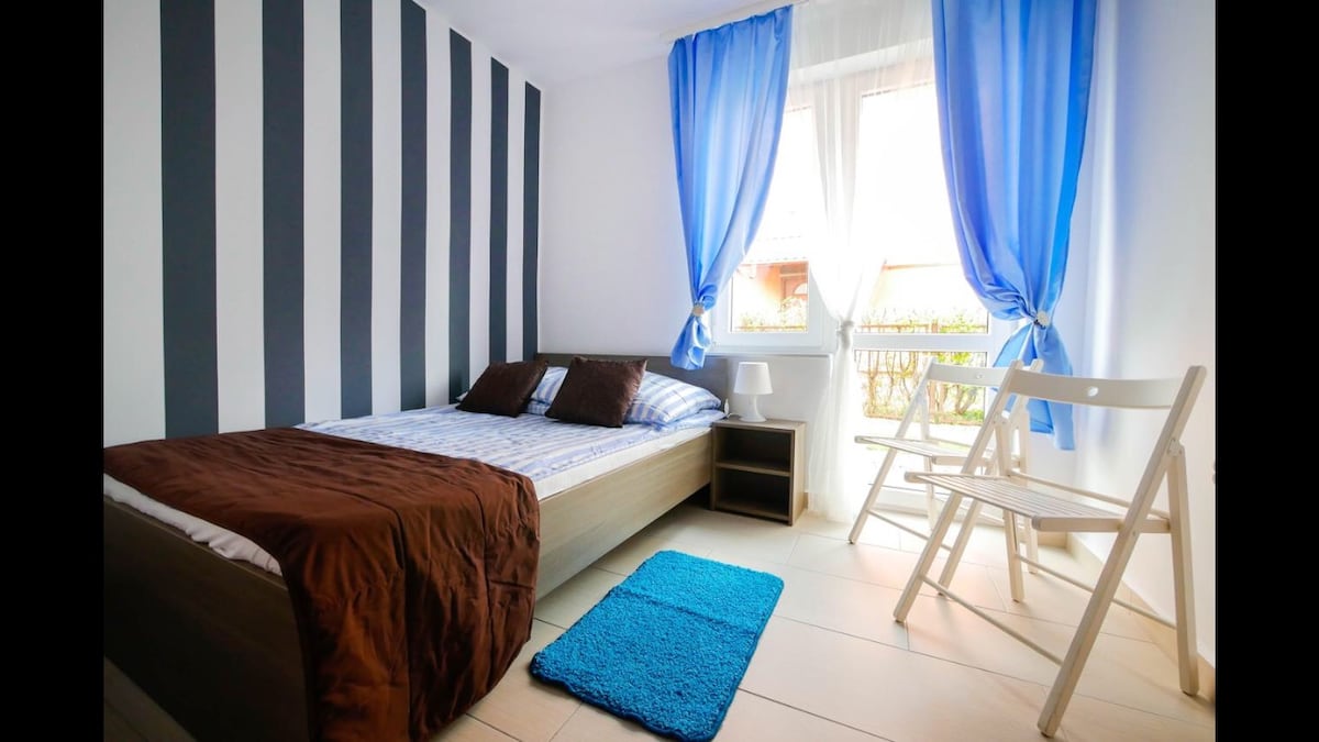 A bright bedroom features striped walls in shades of gray and white. A spacious bed is accompanied by two brown cushions. Nearby, a small chair and a table sit beside a window adorned with blue curtains, allowing natural light to enter the room.
