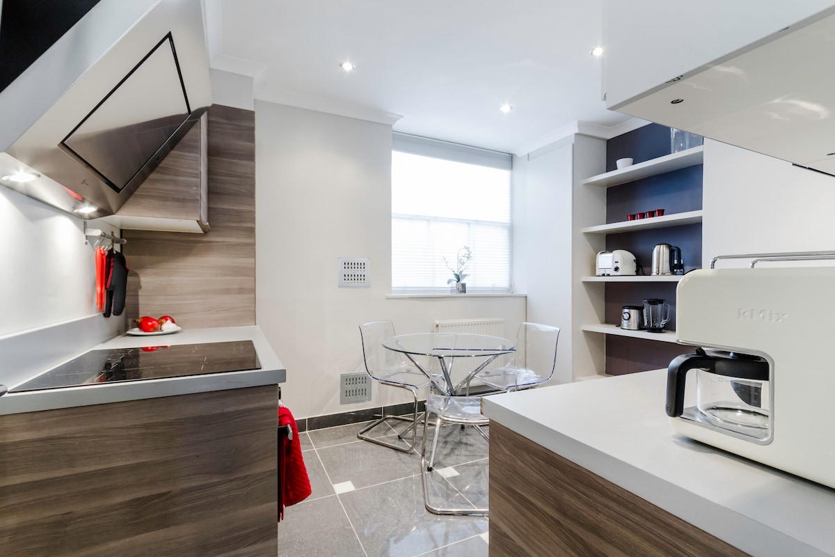 A modern kitchen features sleek cabinetry and high-quality appliances. A glass dining table with transparent chairs is positioned near a bright window. The space is well-lit with recessed lighting, enhancing the crisp design of the grey tiled floor and contrasting textures.