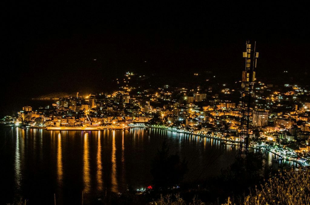 A nighttime view showcases a vibrant seaside town illuminated by city lights reflecting on the water. The gentle glow creates a serene atmosphere, highlighting the varying heights of the buildings along the coastline and the landscape's natural contours.