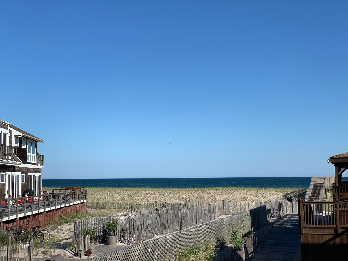 An expansive view of the ocean stretches toward the horizon, framed by a sandy beach and gentle waves. A boardwalk leads through the beach grass, connecting to the nearby property. Clear blue skies enhance the tranquil seaside environment.