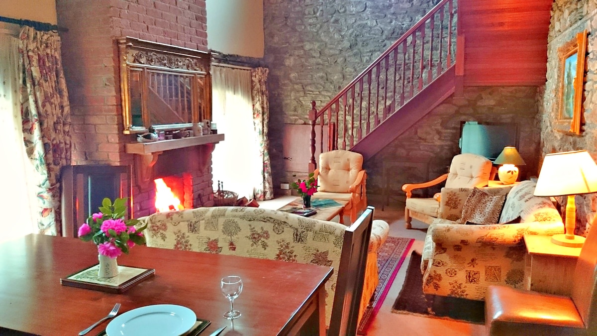 The inviting living area features a blend of stone and wood elements, creating a cozy atmosphere. A fireplace is centered in the room, with soft seating arranged around it. A dining table with decorative flowers is visible, complemented by warm lighting from nearby lamps.