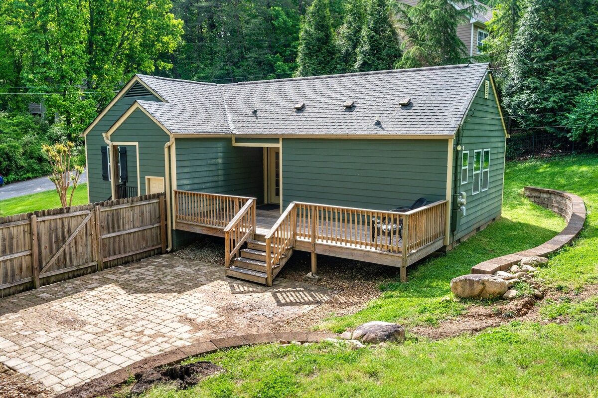 The exterior of a green house is shown, featuring a wooden deck with steps leading down to a stone-paved patio. A fenced area surrounds the yard, with trees and greenery visible in the background, creating a serene outdoor space.