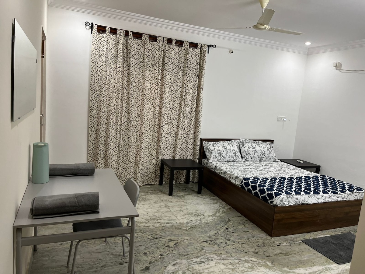 A clean and simple room features a spacious bed with patterned bedding and a wooden bed frame. A small table and two chairs are positioned nearby, while a light-colored curtain adds texture to the window. The floor displays a glossy marble finish.