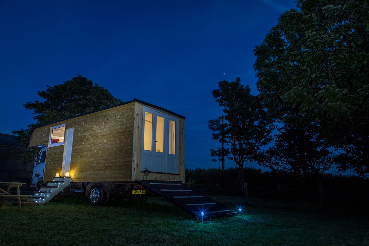 The converted horse lorry is shown at night, with warm light glowing from its windows. Soft lights illuminate the steps leading up to the entrance, while surrounding trees are gently outlined against the evening sky.