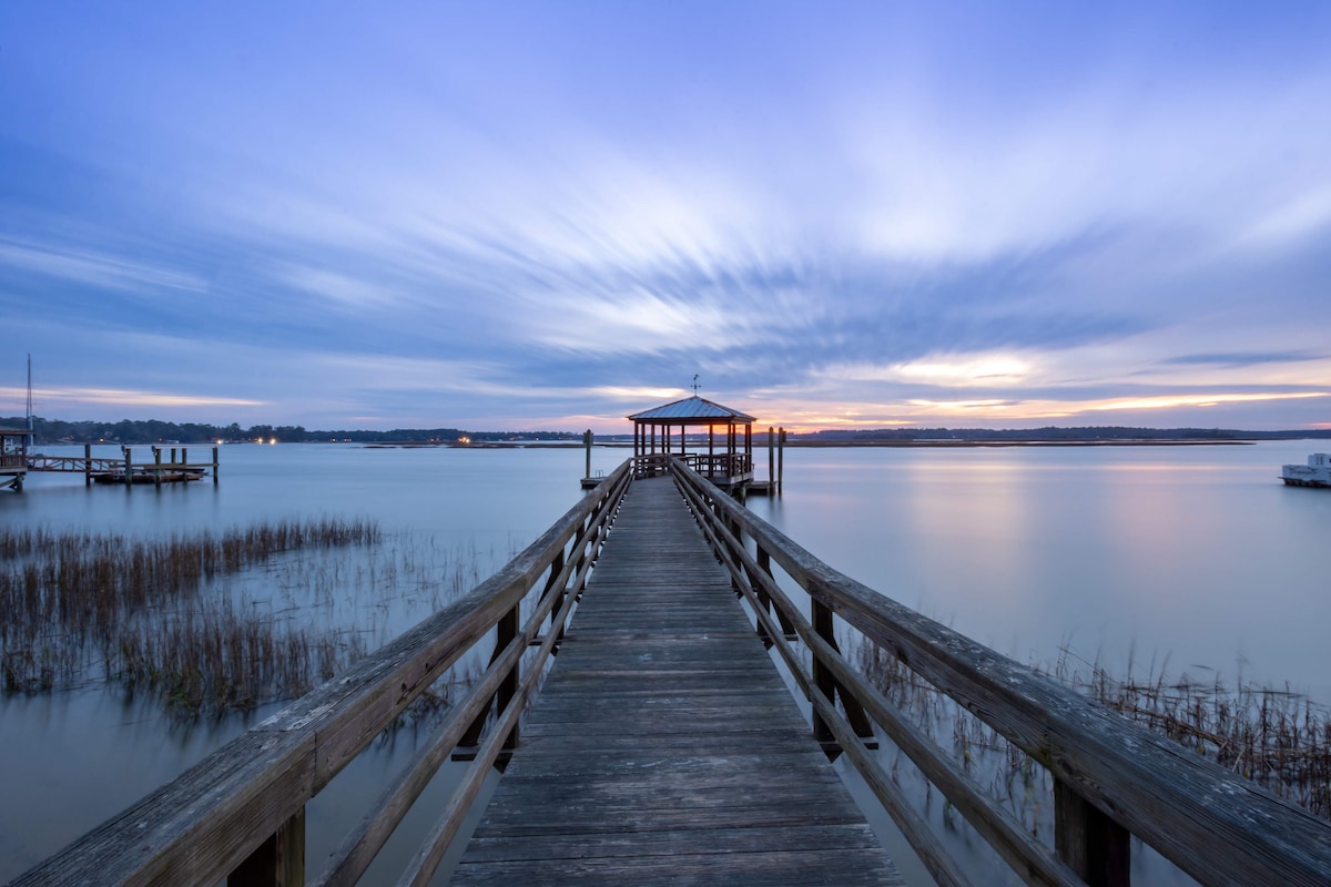 A long wooden dock extends over calm waters, leading to a gazebo under a gently illuminated sky. The horizon features soft pastel colors, while subtle reflections shimmer on the water's surface. Tall grass borders the dock, enhancing the serene riverside setting.