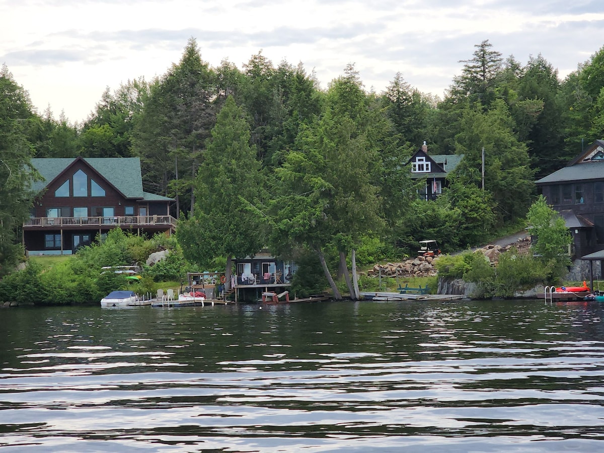 A serene lakeside view depicts a collection of cabins nestled among lush trees. Gentle ripples reflect the greenery and sky on Upper Saranac Lake's surface. A private dock and outdoor seating area, along with various watercraft, enhance the tranquil atmosphere of this natural setting.