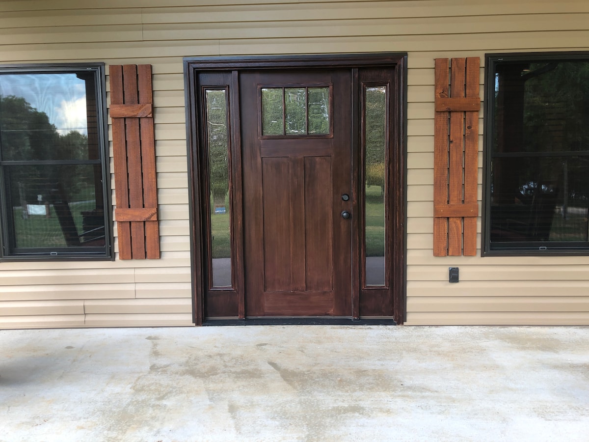 A rustic wooden front door is framed by two decorative shutters, presenting a sturdy and welcoming entrance. Large windows on either side allow natural light to brighten the exterior. A concrete porch leads up to the door, enhancing accessibility.