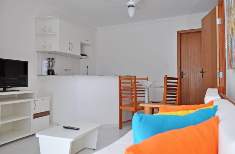 A bright living area is featured, showcasing a comfortable sofa adorned with colorful cushions. A flat-screen television is positioned on a media console. The open kitchenette is visible, equipped with essential appliances, while a dining table is set for four with wooden chairs.
