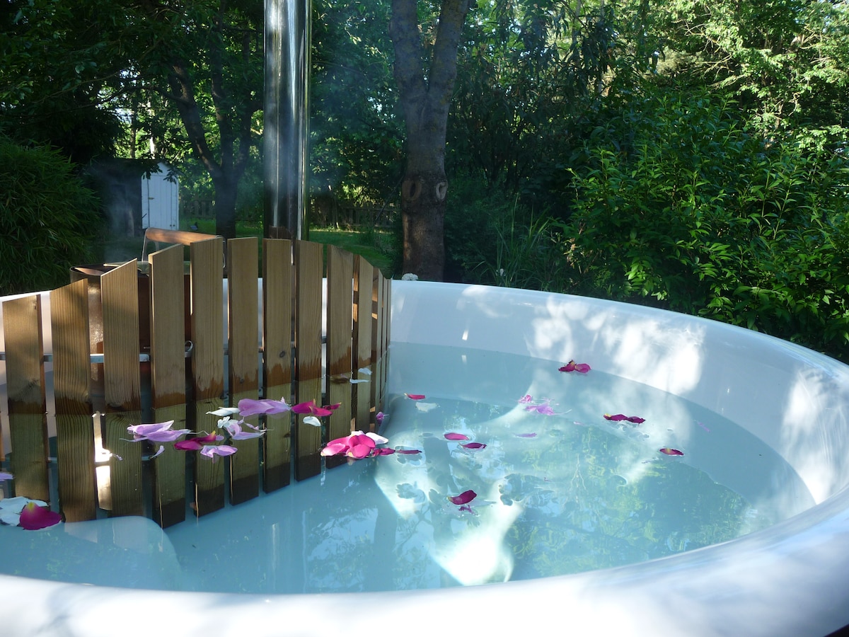 A hot tub is surrounded by a wooden privacy screen and natural greenery. The water is serene, featuring floating pink and white petals that add a gentle touch of color to the tranquil setting.