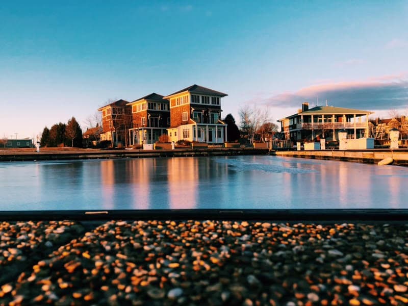 A serene view of three elegantly designed homes situated along a frozen lakeside. The structures feature varied color palettes and architectural styles, reflecting natural light from the water's surface. A clear sky complements the tranquil scene, enhancing the peaceful atmosphere of the location.
