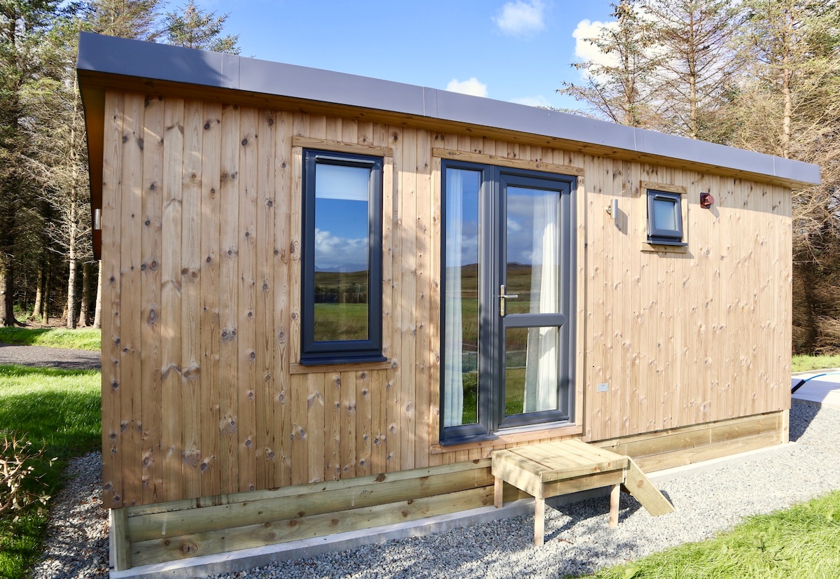 The modern cabin exterior features a wooden façade with large windows, allowing natural light to enter. A small deck leads to the front door, and a gravel path enhances accessibility. Green grass surrounds the cabin, while trees provide a natural backdrop.