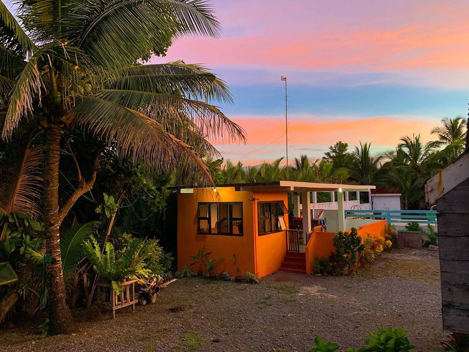 A colorful building with bright orange walls is surrounded by lush greenery and tall palm trees. The evening sky features soft pink and orange hues, creating a serene backdrop. A gravel path leads to the entrance, offering a welcoming feel to the tranquil environment.