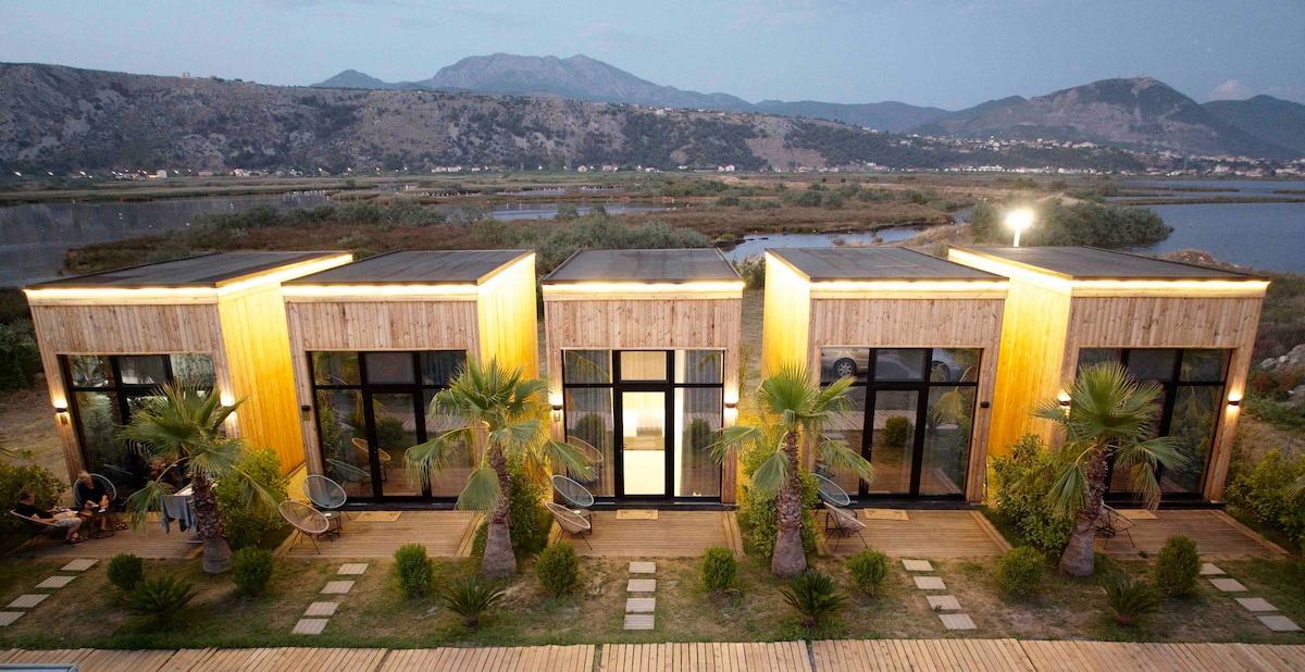 A row of modern, wooden structures is set against a backdrop of rolling mountains. Large glass windows reflect the surrounding nature, while palm trees and manicured greenery line the pathway. Outdoor seating areas invite relaxation in a serene environment.