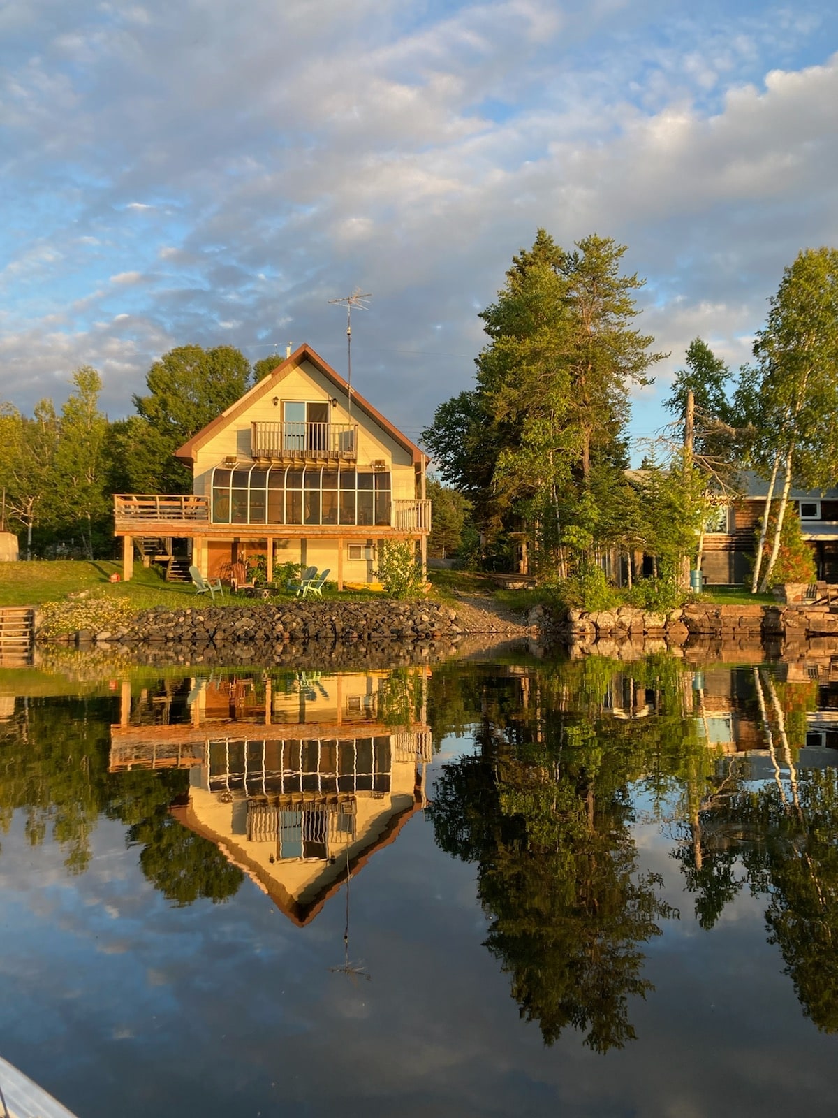 A lakeside home is depicted, featuring a peaked roof and large windows reflecting in the calm water. A spacious deck is visible, surrounded by greenery and mature trees. The peaceful setting is enhanced by a clear sky and soft clouds, creating a serene atmosphere.