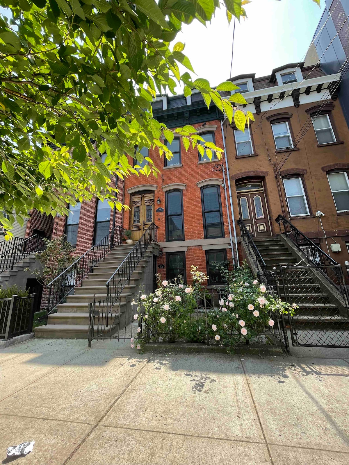 A classic brownstone exterior is depicted, showcasing a brick façade with prominent steps leading to the entrance. Ornamental iron railings flanking the stairs add character, while lush greenery and blooming roses are visible in the foreground, creating a welcoming atmosphere.