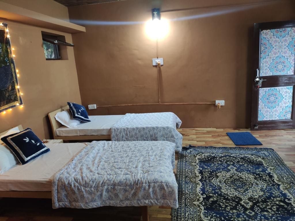 A cozy room features two spacious beds, each dressed with soft blankets and decorative cushions. A patterned rug lies on the wooden floor, adding warmth. The walls are painted in a warm tone, and a soft light fixture provides illumination, enhancing the room's tranquil ambiance.