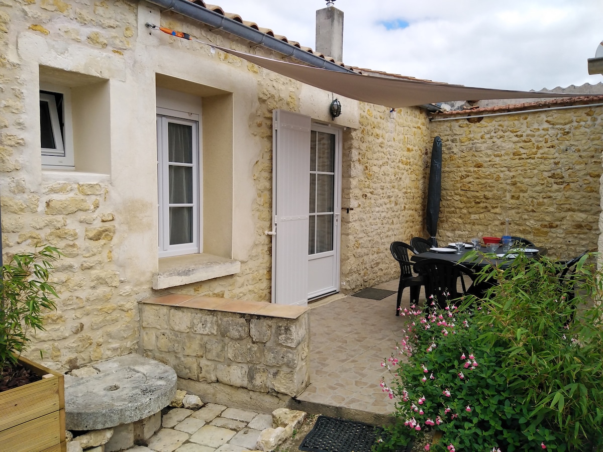 The outdoor terrace features a stone wall and is furnished with a dining table and black chairs. A large umbrella provides shade, while a small flowerbed adds a touch of greenery. The entrance to the accommodation is highlighted by a white door and window.