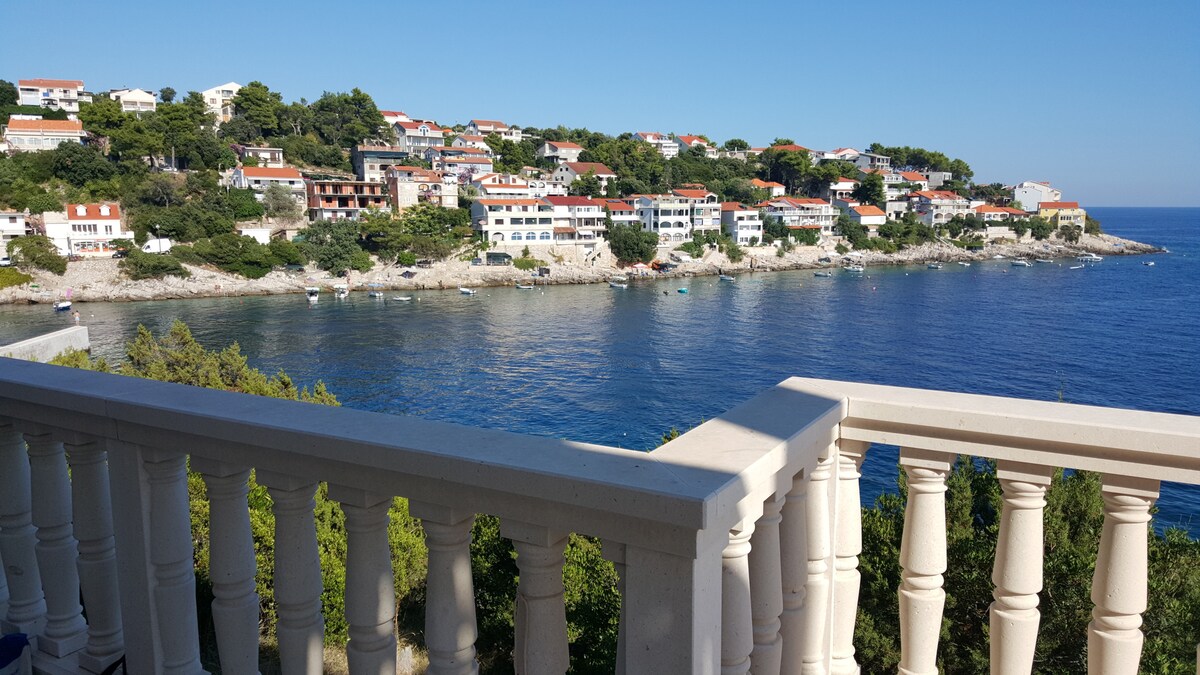 A coastal view is presented from a balcony featuring balusters, overlooking a serene bay. Colorful houses line the hillside, complemented by lush greenery. The calm sea reflects the clear blue sky, creating a peaceful atmosphere in the landscape.