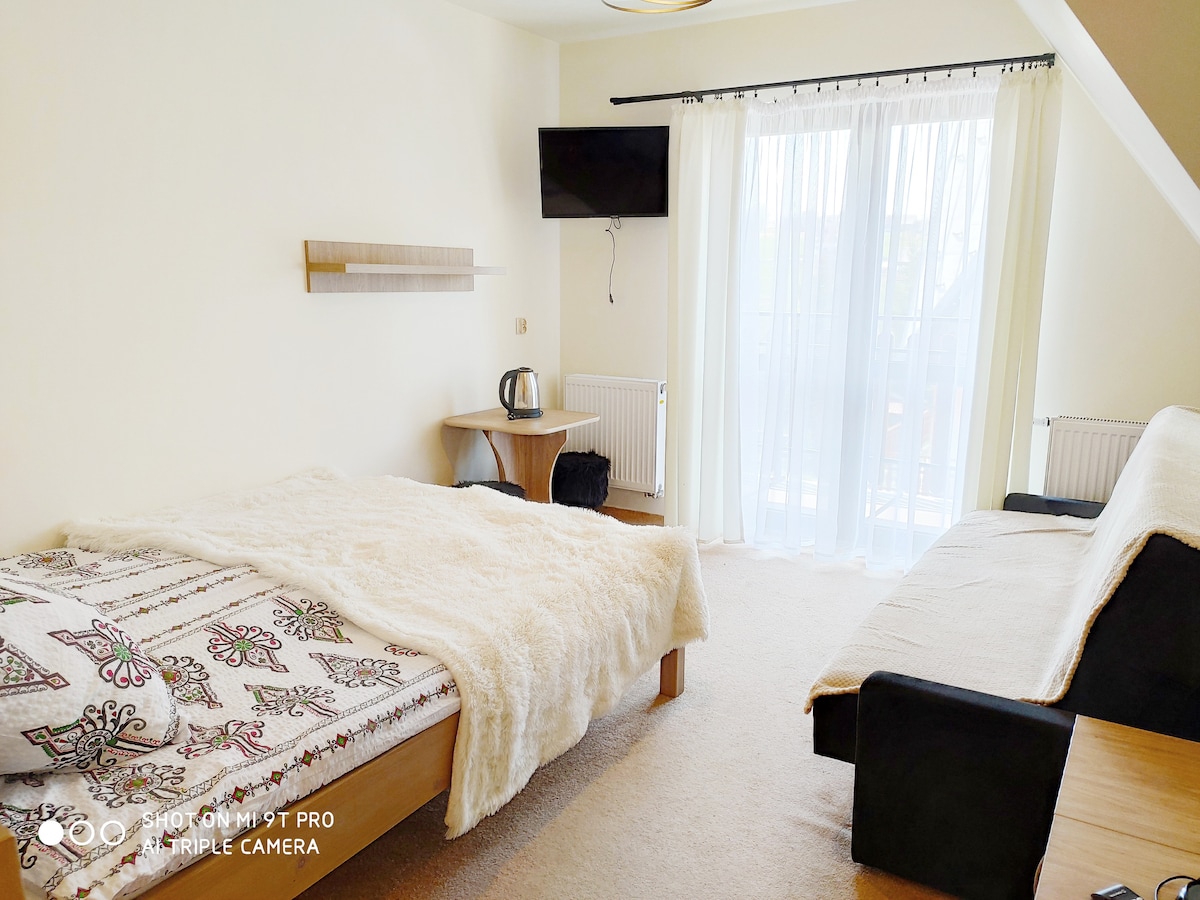 A bright room features a spacious bed covered with a cozy blanket, alongside a sofa and a small table. A television is mounted on the wall, and natural light floods in through the large glass doors leading to a private balcony.