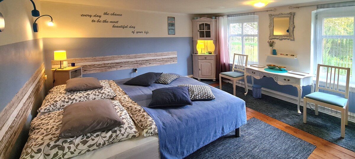 A spacious sleeping area features a large bed with multiple pillows and a blend of patterned linens. Two large windows allow natural light to fill the room, complemented by soft blue walls and wooden accents. A small desk and chair are poised near a decorative mirror.