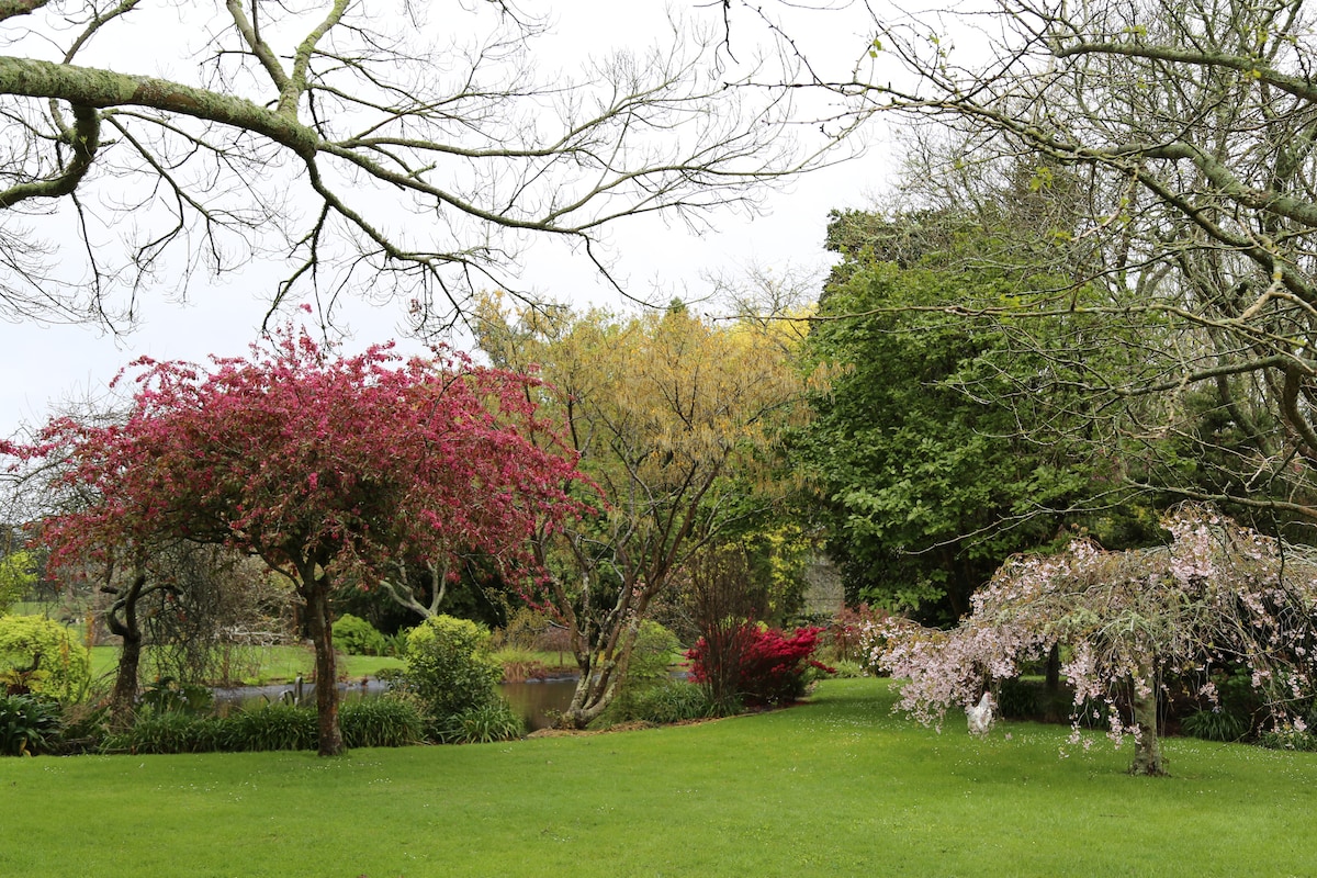 The image displays a serene garden landscape featuring a lush green lawn surrounded by various trees. Vibrant blossoms appear in shades of pink and red, contrasting with the greenery. A tranquil pond is partially visible in the background, enhancing the peaceful atmosphere of the setting.
