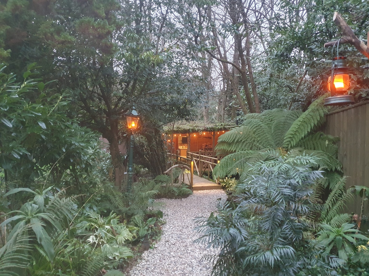 A gravel path meanders through lush greenery, leading to a warmly lit cabin nestled among trees. Lanterns provide soft illumination, highlighting the dense foliage and inviting visitors toward the entrance of the cabin.
