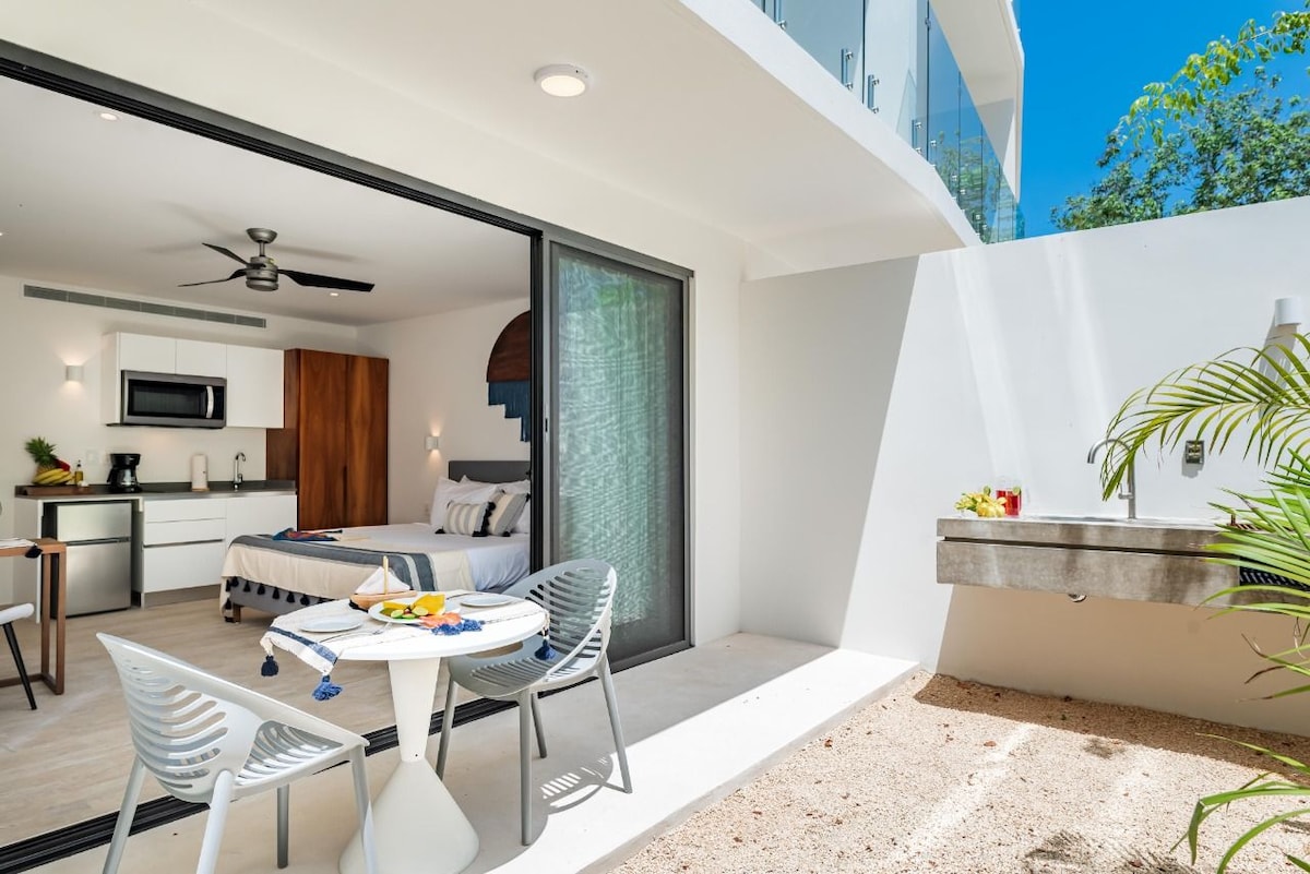 An open space features a modern kitchen area and a cozy sleeping area with a king bed. A small dining table is accompanied by four chairs. Natural light fills the room from large glass doors that open to a private outdoor area with a washbasin.