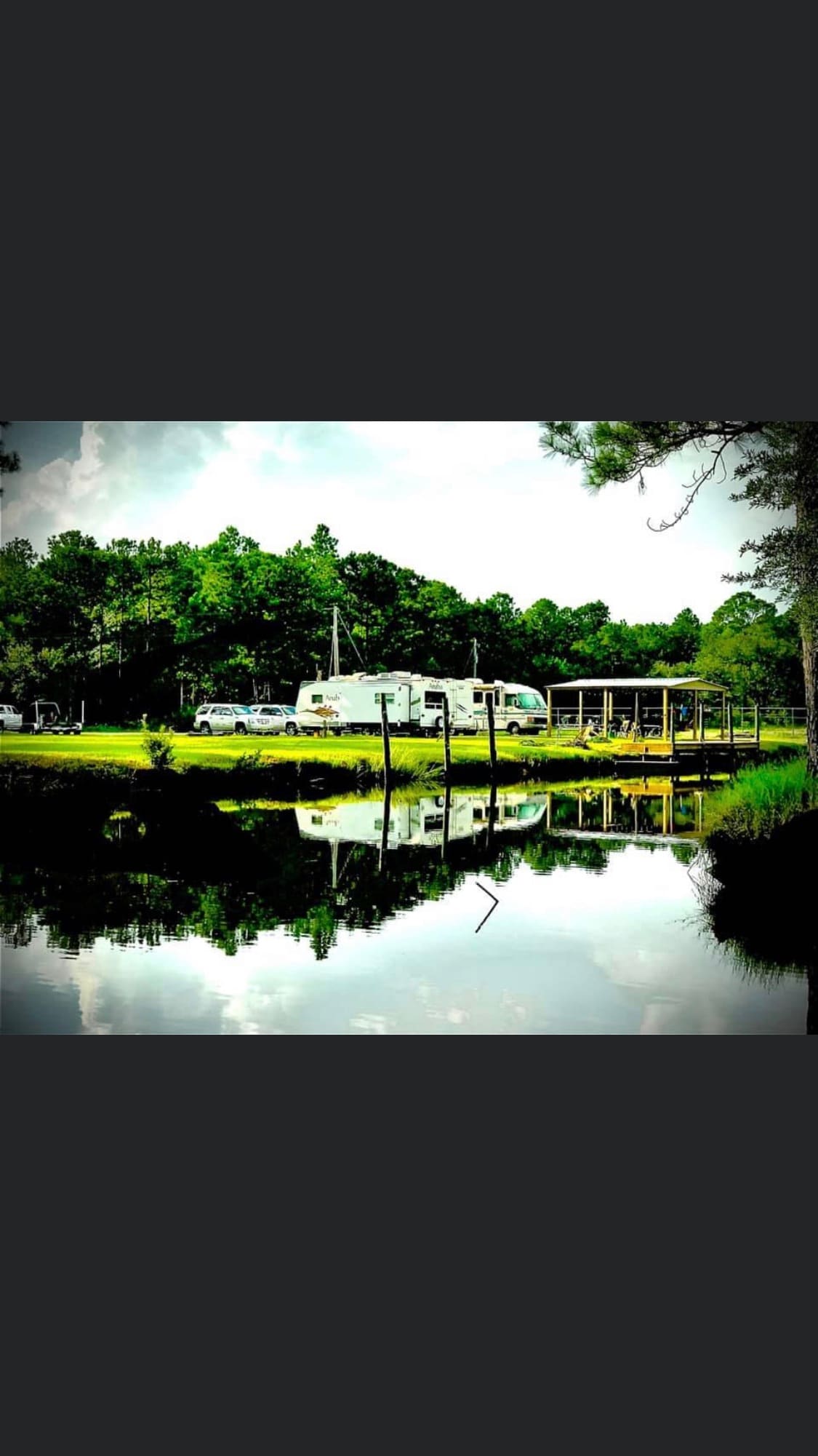 The image shows three RVs parked near a tranquil body of water, reflecting the surrounding lush greenery and pavilion. A covered structure is visible in the background, providing shade, while tall trees frame the serene scene, creating a secluded ambiance.