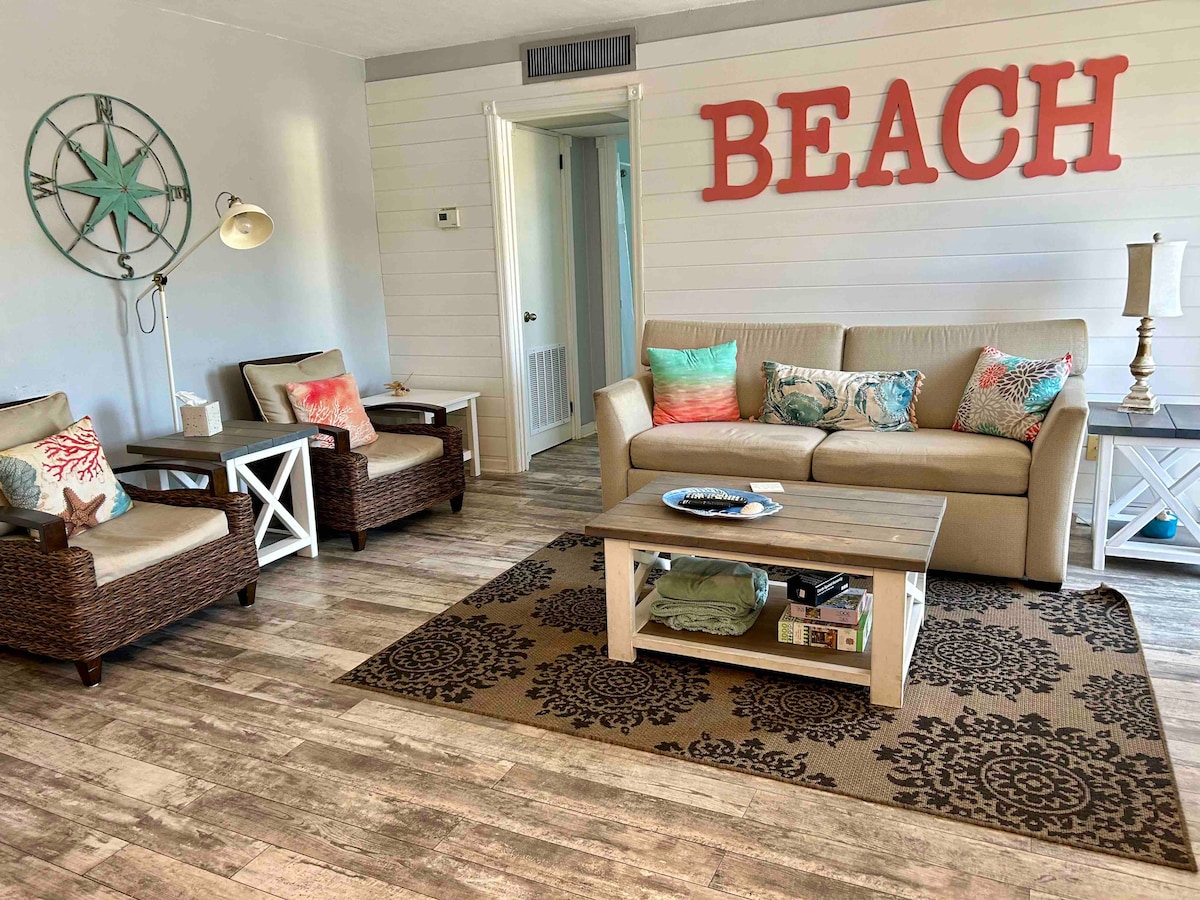 A comfortable living area is highlighted by a tan sofa with decorative pillows, accompanied by two wicker chairs. A wooden coffee table with items displayed sits on a patterned rug. The word 'BEACH' is prominently featured on the wall, enhancing the coastal theme.