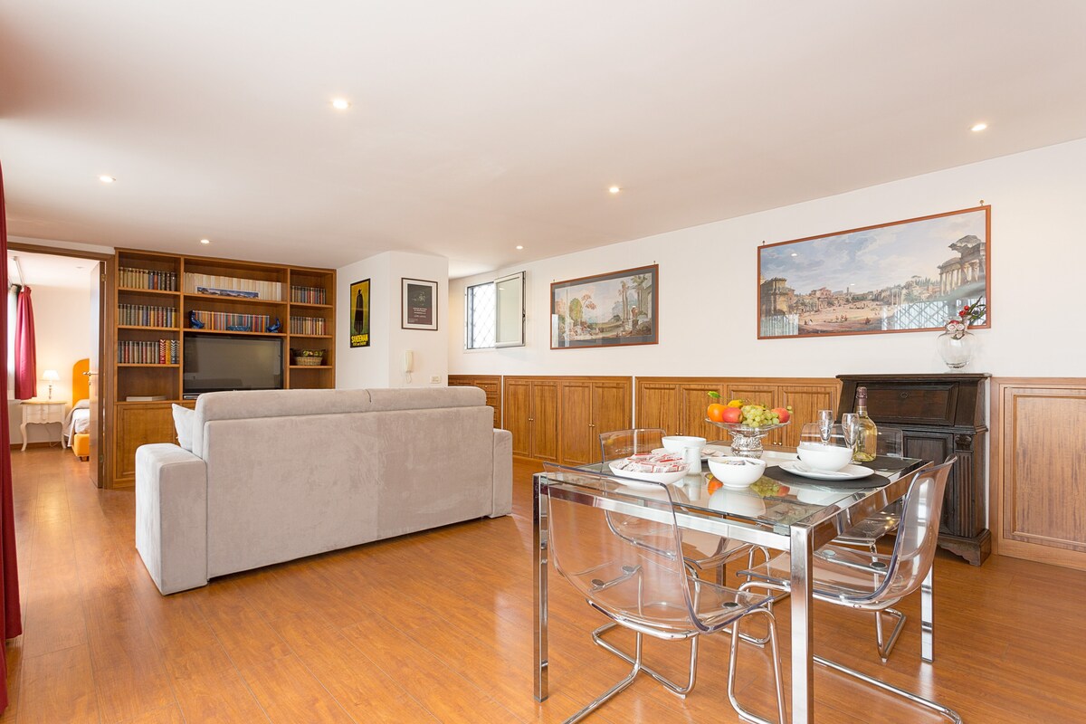 A spacious living area features a light wall design and warm wooden accents. A modern glass dining table is set with tableware and food, while a comfortable sofa is positioned nearby. A bookshelf and artwork decorate the space, creating an inviting atmosphere.