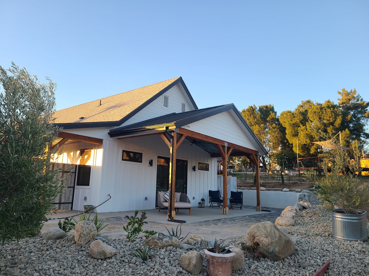 A modern corner villa is presented with a combination of white siding and a dark roof. The spacious outdoor area features a stone patio, accented by native plants and decorative boulders, creating a serene atmosphere under a clear blue sky.