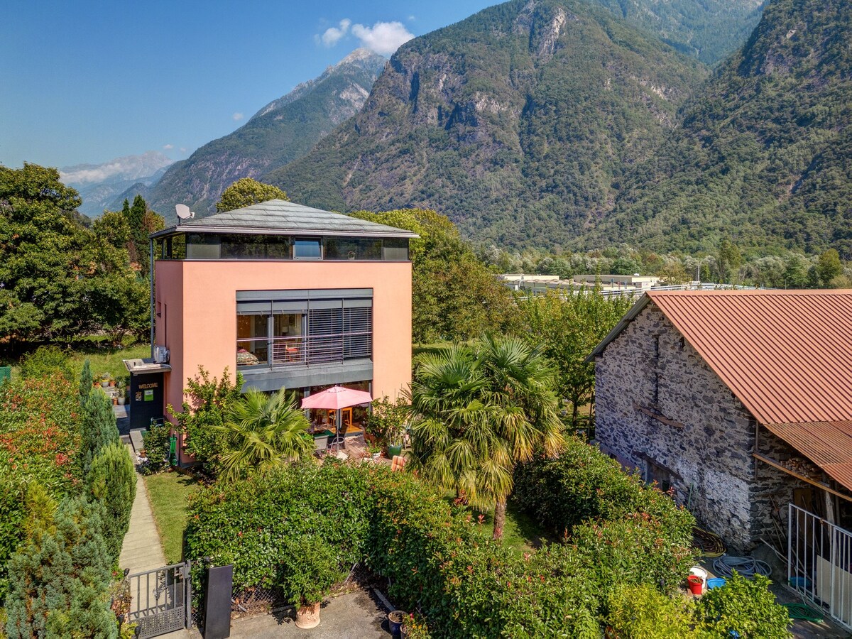 The image captures a modern, two-story building nestled among lush greenery, with mountains rising in the background. Outdoor seating areas are visible, shaded by a large umbrella. A separate stone structure nearby adds rustic charm to the serene landscape.