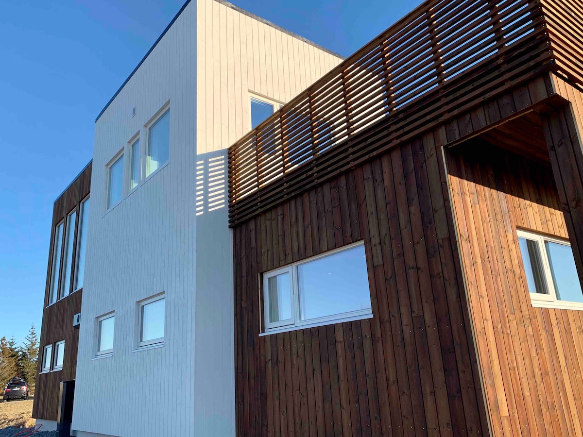 The modern house showcases a mix of white and wood paneling, highlighting its contemporary design. Large windows provide ample natural light, and a wraparound balcony is visible on the upper level, enhancing the architectural features.