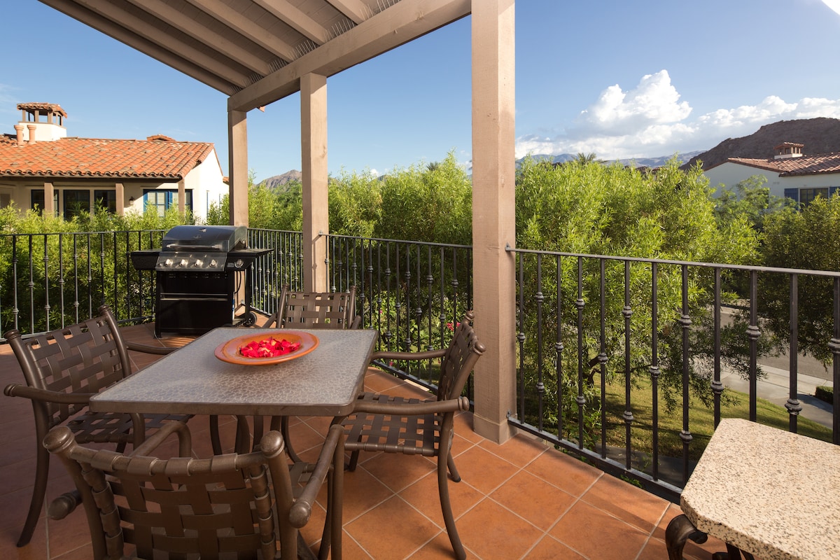 A spacious balcony is highlighted by a dining table and four chairs, complementing a grill in the corner. Lush greenery surrounds the space, offering privacy and a view of the mountains. A clear sky and distant buildings are visible in the background.