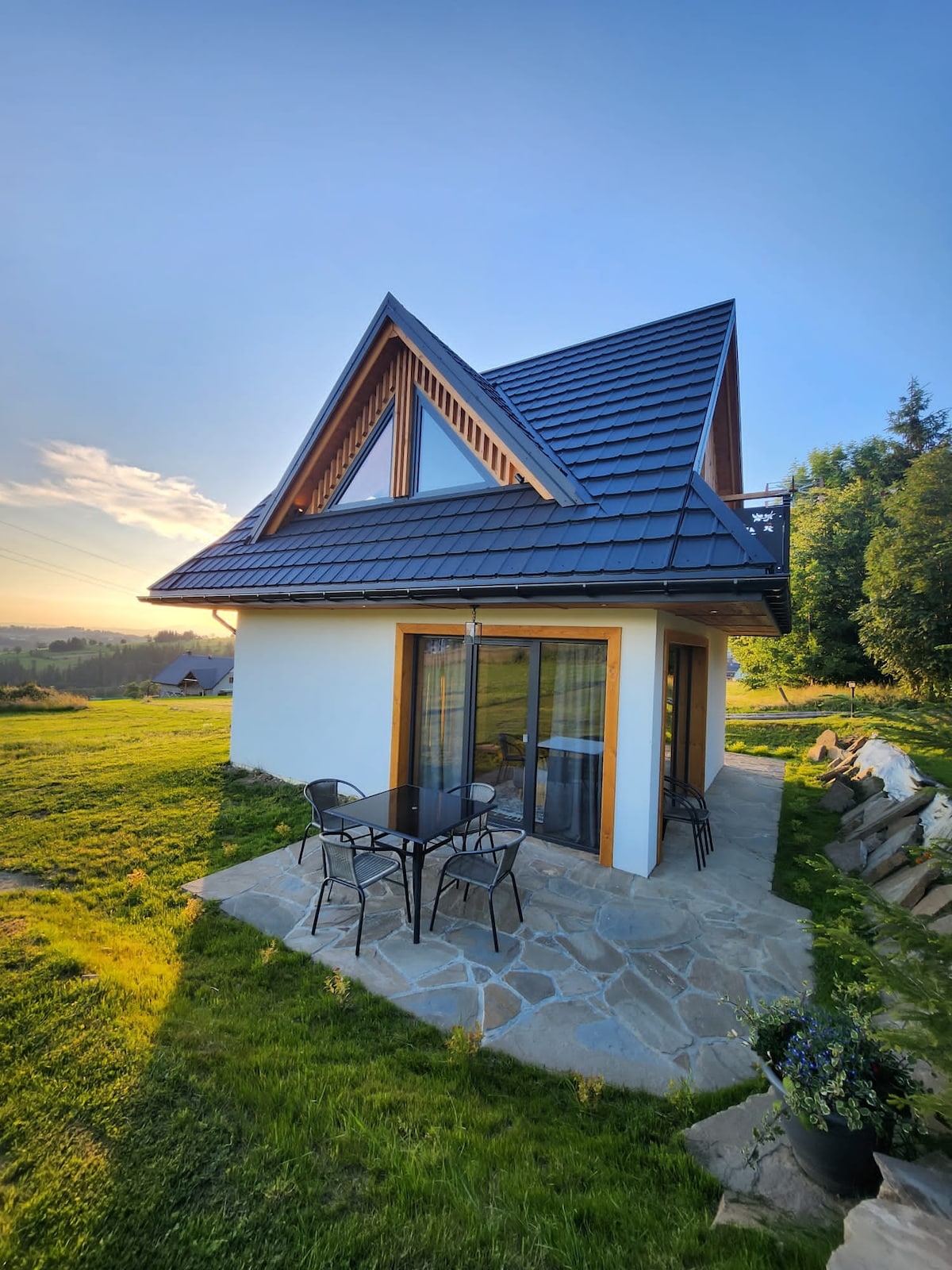 The exterior of a contemporary chalet is displayed, featuring a sloped roof and large glass doors. A stone patio is furnished with a small table and chairs, surrounded by green grass and landscaping. The backdrop includes rolling hills and a partially clear sky.