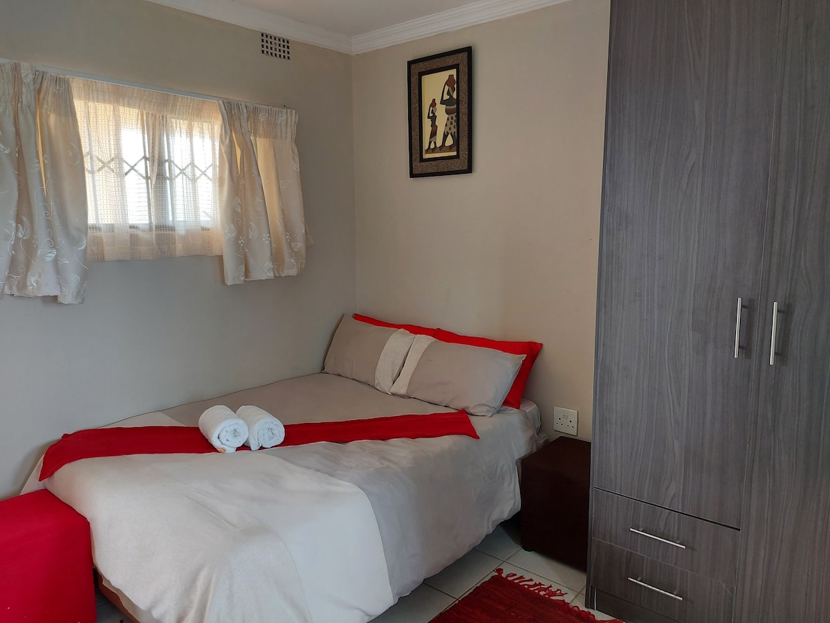 A cozy bedroom features a spacious bed adorned with neutral bedding and vibrant red accents. Fresh towels are neatly arranged on the bed. Natural light enters through a curtained window, and a wardrobe stands against the wall, offering ample storage.