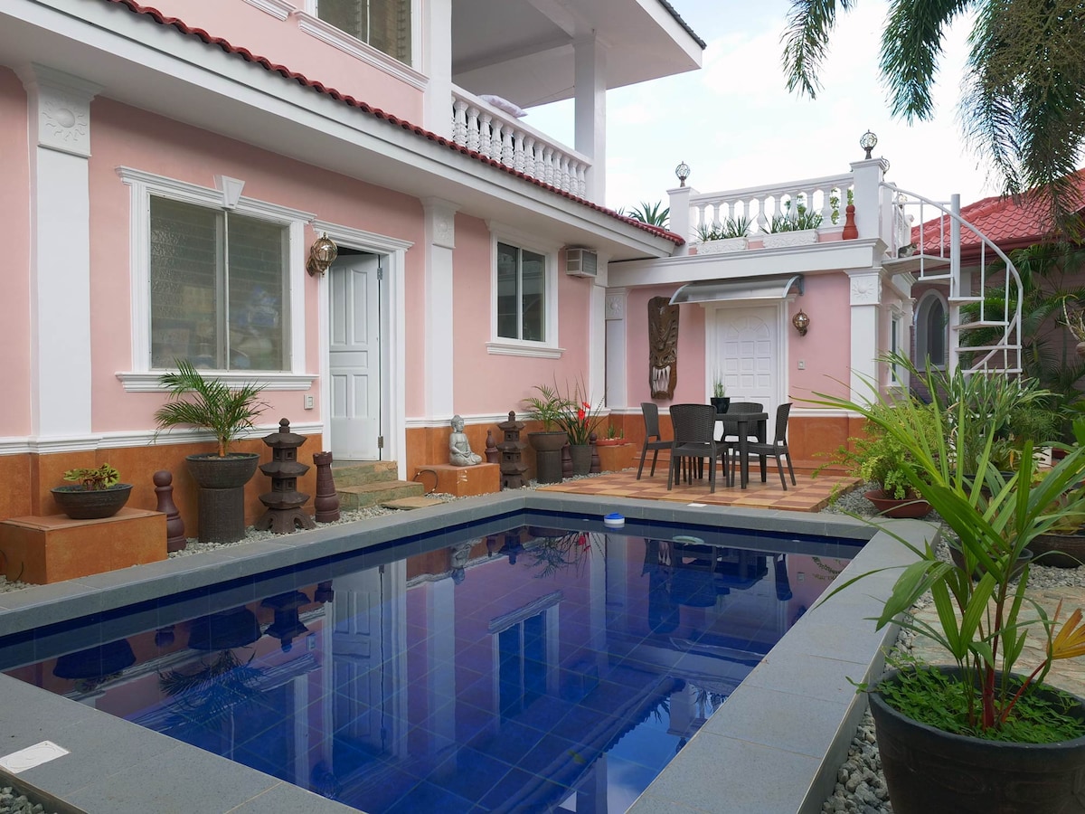 An exterior view showcases a well-maintained private pool surrounded by lush greenery. The patio features an outdoor dining set and ample stone pathways, creating a welcoming outdoor space. The pink facade of the house adds a charming touch to the serene garden setting.